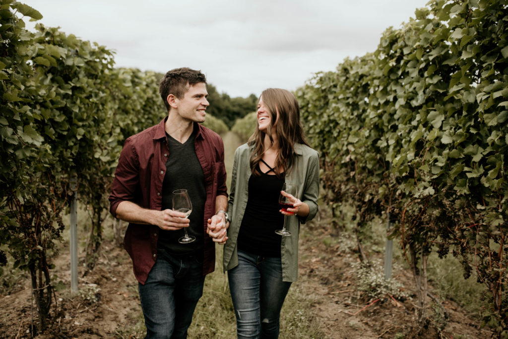 couple walking through vineyard holding wine glasses. woman wearing olive green. man wearing maroon red. engagement portrait photos