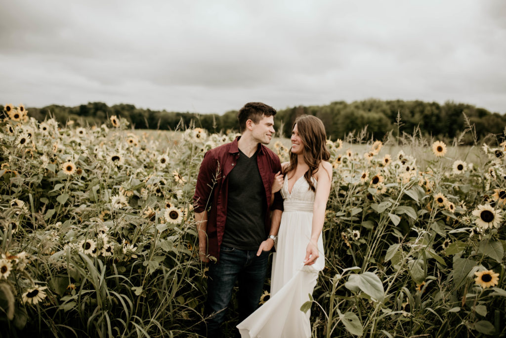 couple standing in sunflower field. man wearing maroon red shirt, woman wearing long white lacy boho dress. engagement portrait photo.