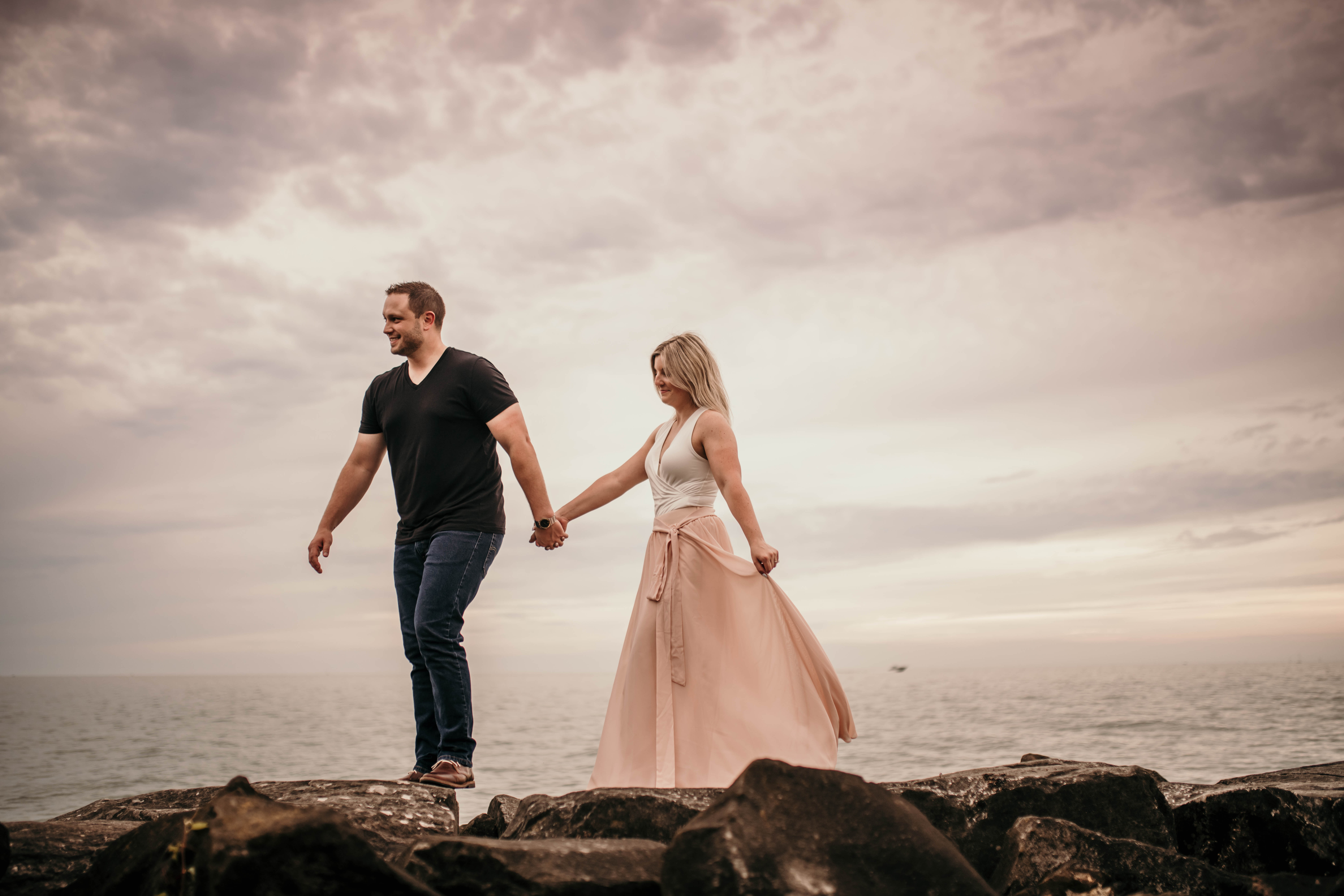 couple walking on rocks by water. woman wearing long flowy boho pink skirt. man wearing black simple shirt. warm tone engagement portrait photo.