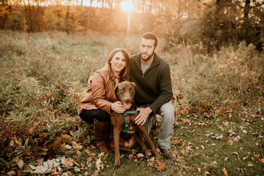 couple with dog sitting in field. fall leaves on ground. brown chocolate lab dog. warm tone fall engagement photo portrait