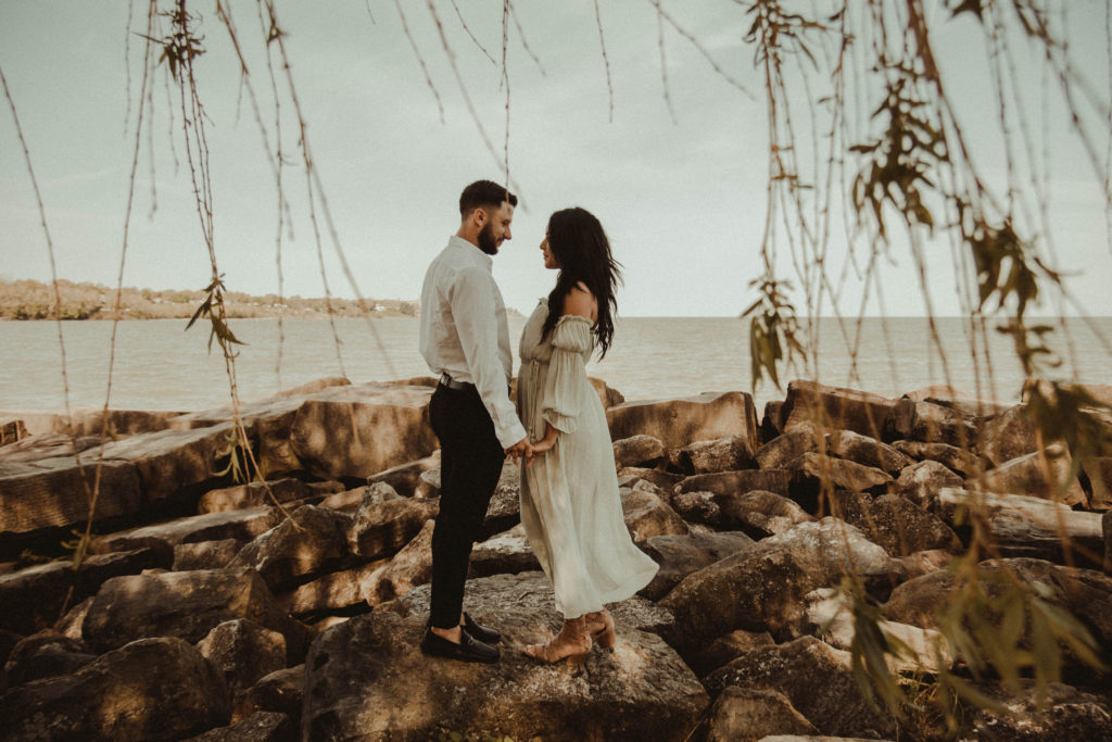 couple standing on rocks by water with leaves hanging in front. engagement portrait photos