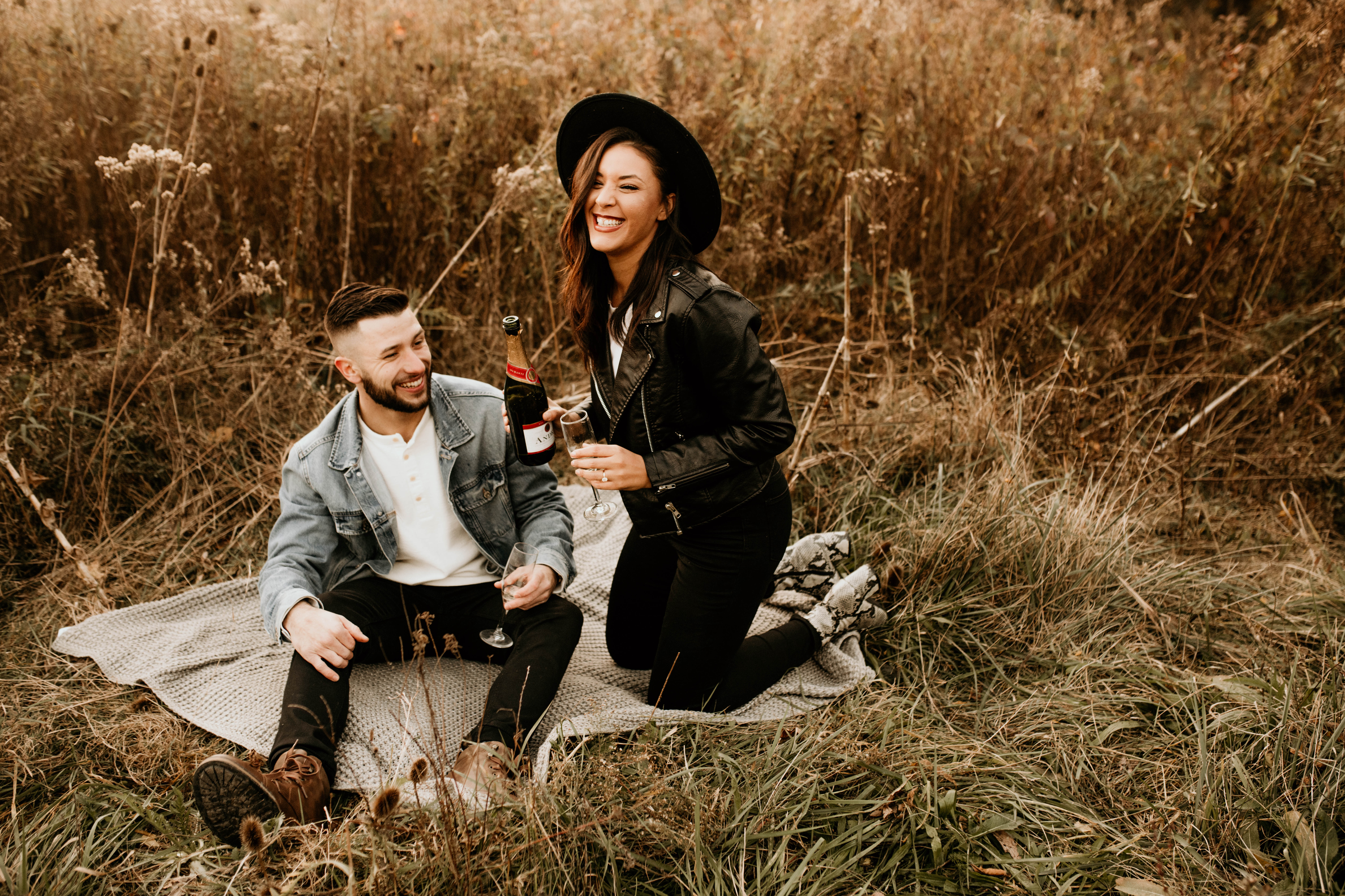 Edgy Engagement Style, Man wearing jean jacket and Woman black leather jacket and hat sitting on blanket with wine.