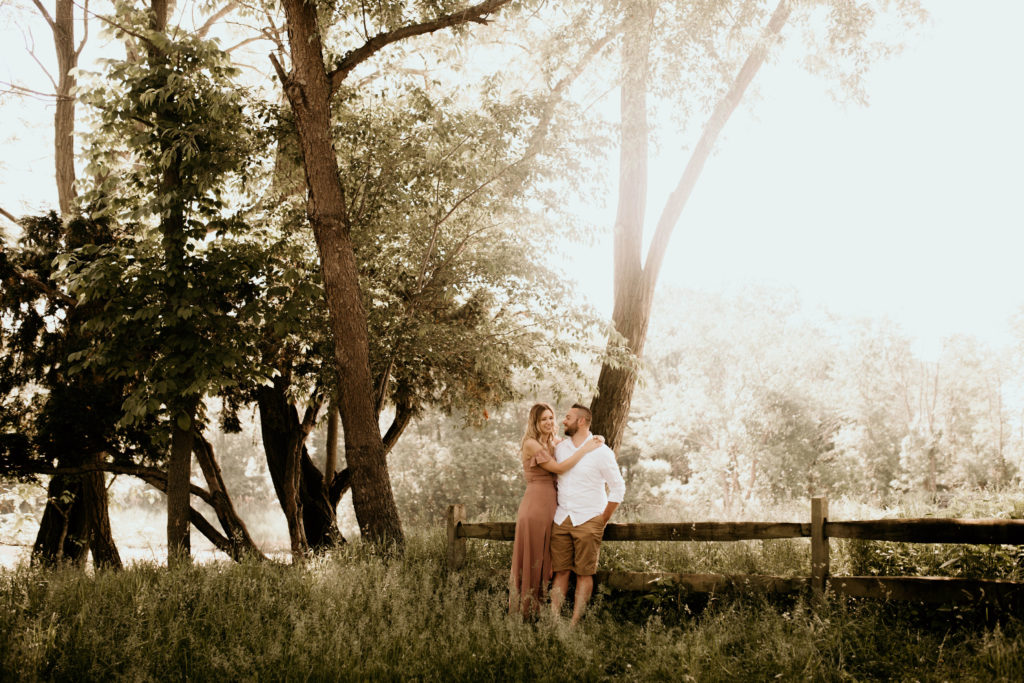 couple leaning on fence in field with trees in the background. engagement portrait photo