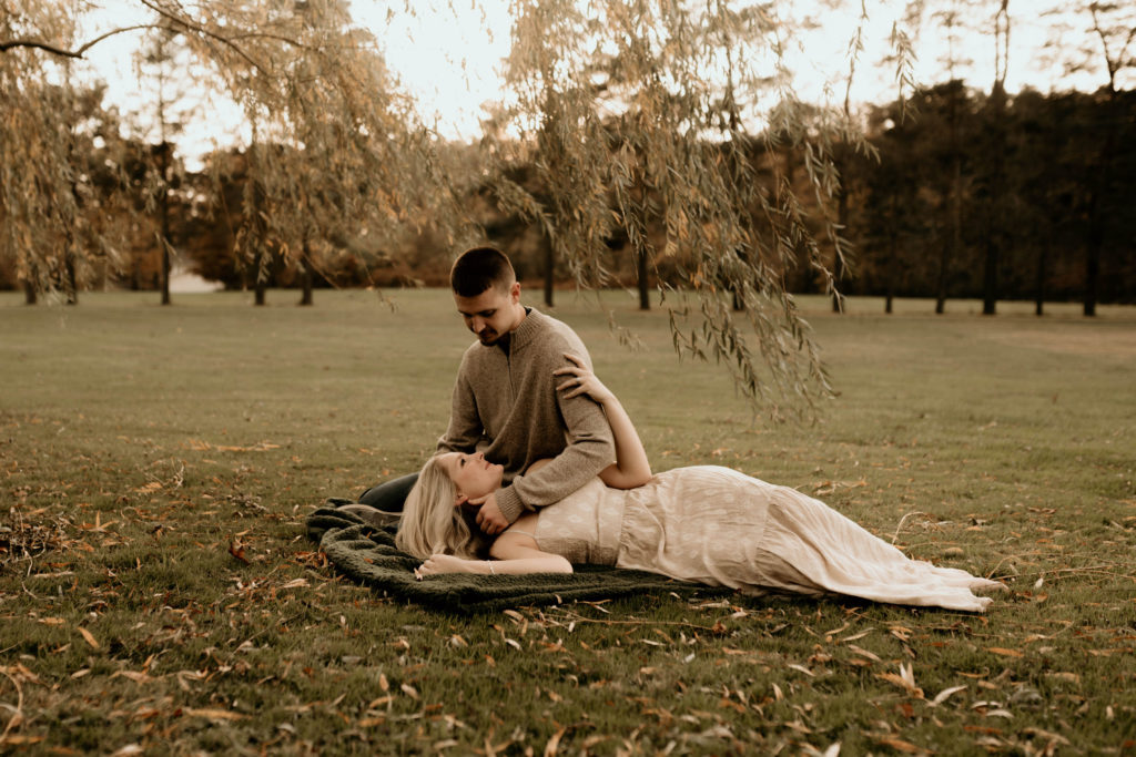 couple sitting in field. leaves in foreground. woman in white boho lace dress laying on ground. man in grey sweater. romantic warm tone engagement portrait photo