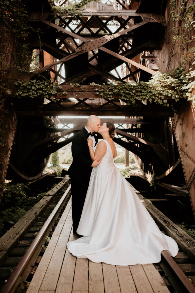 bride and groom kissing on industrial train bridge with ivy
