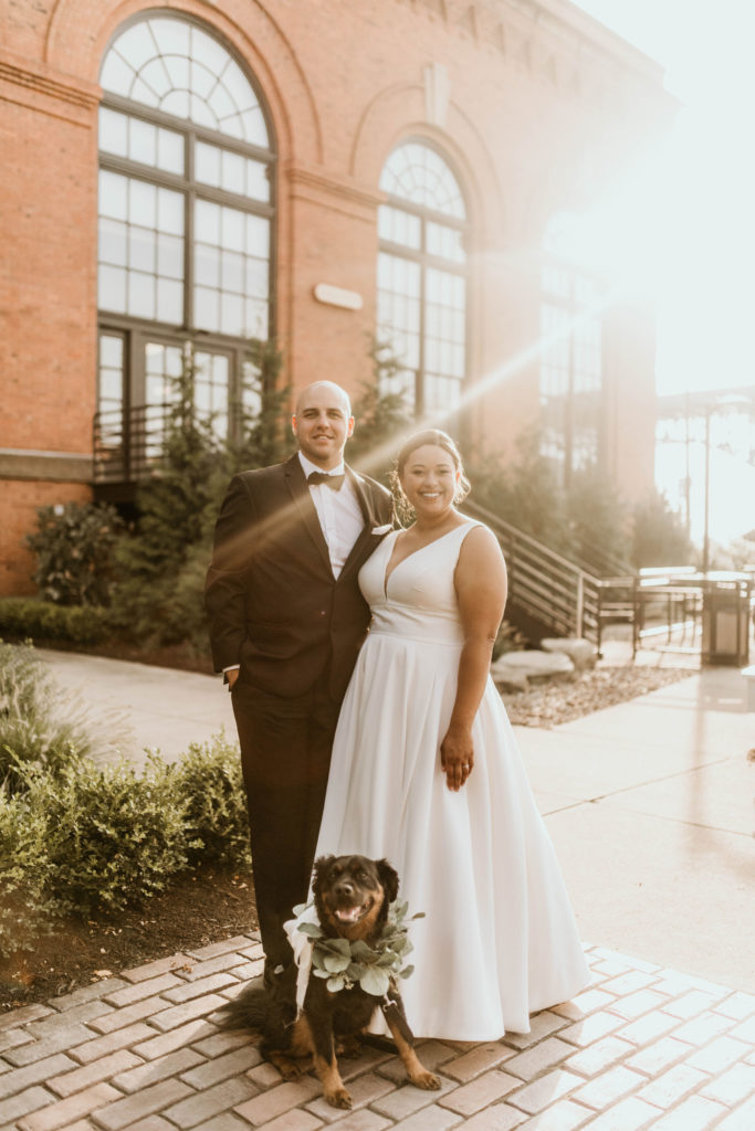 bride, groom, and dog standing together in front of brick building with sun flare
