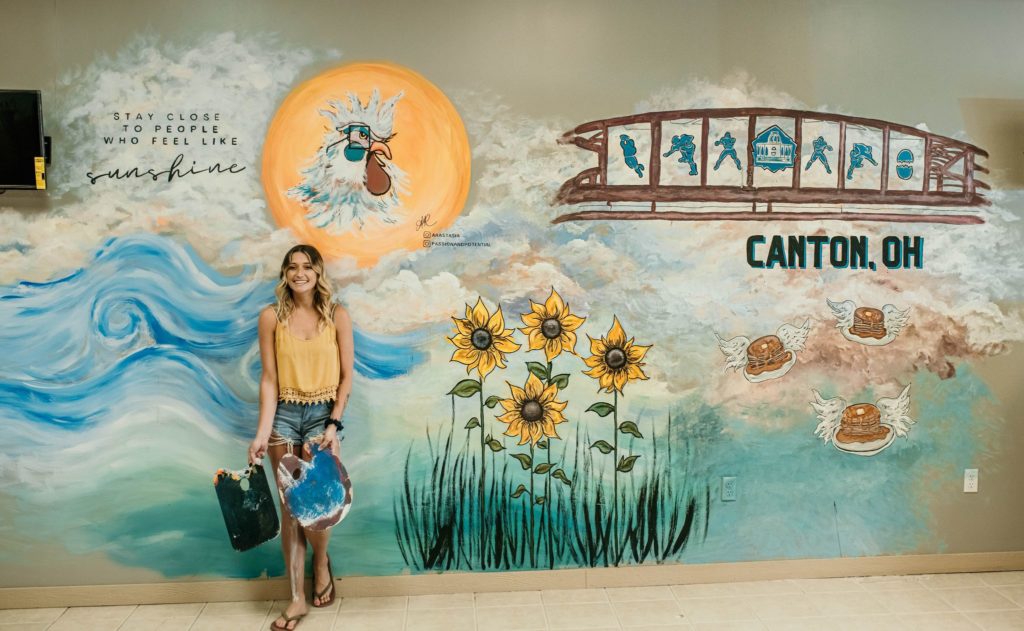 Arastasia standing in front of her mural at the cracked egg in canton ohio. The mural contains clouds, flying pancackes, sunflowers, sun, and a chicken